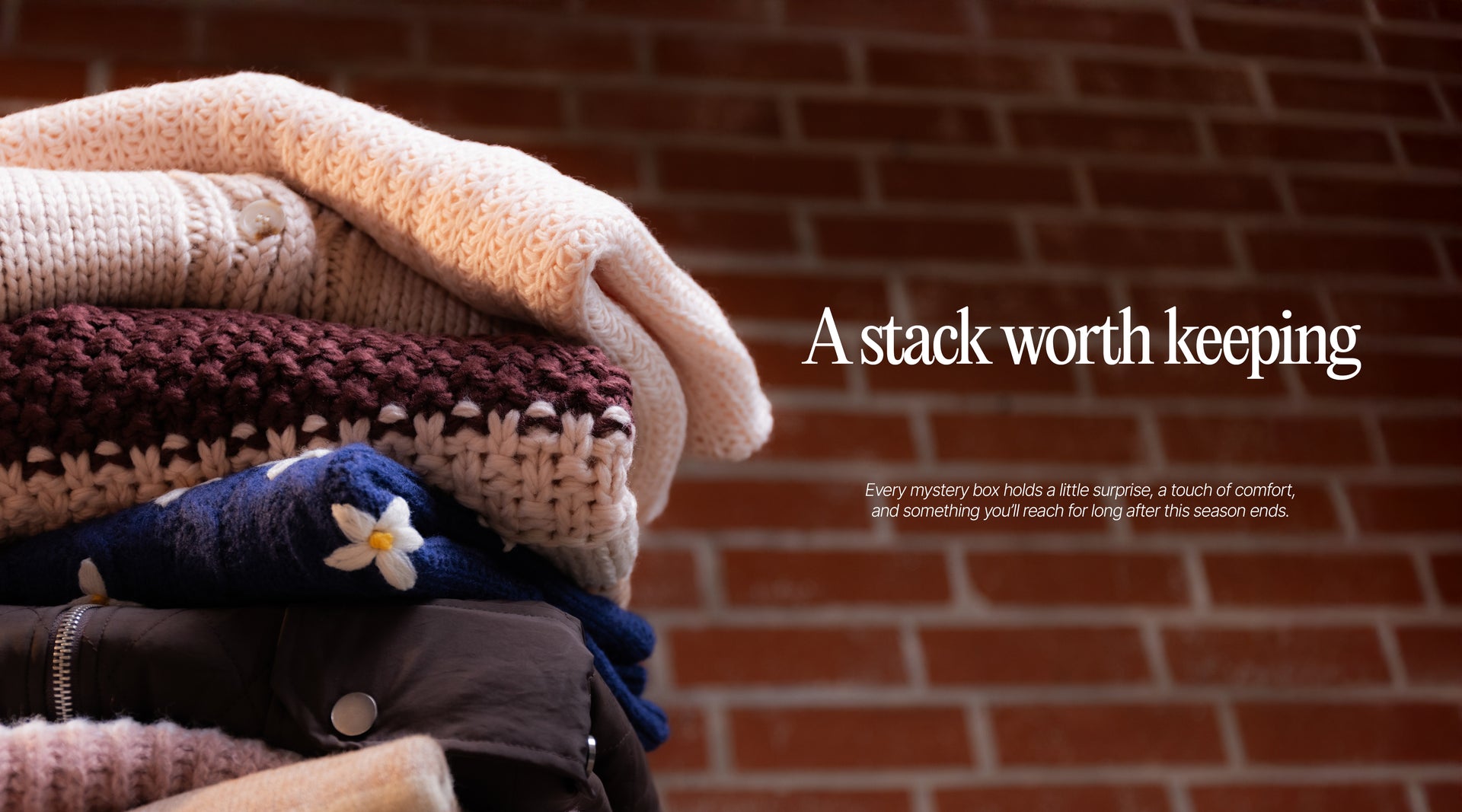 Stack of folded clothes with a brick wall background and text overlay.Next to this is Main text that says 'A stack worth keeping' and subtext underneath that says 'Every mystery box holds a little surprise, a touch of comfort, and something you'll reach for long after this season ends."